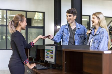 Smiling hotel receptionist handing a passport back to a young couple at the front desk, representing a friendly check-in experience and review.